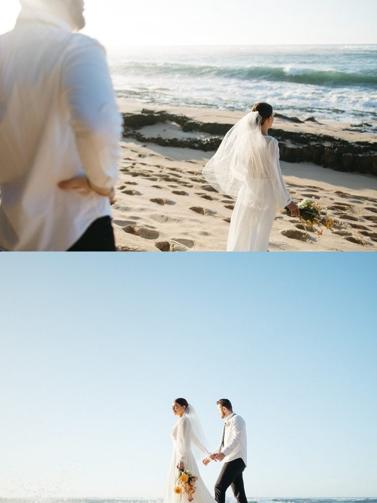 groom is looking at his wife, her back is turned to him. the coupple is walking along the beach holding hands