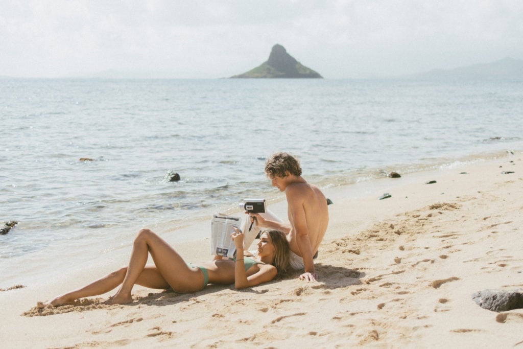 couple is laying on the beach. girl is reading and the boy is doing film photography