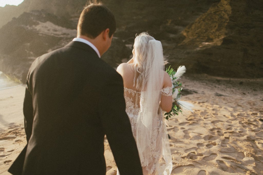 groom is holding the train of the brides dress as she walks along the beach