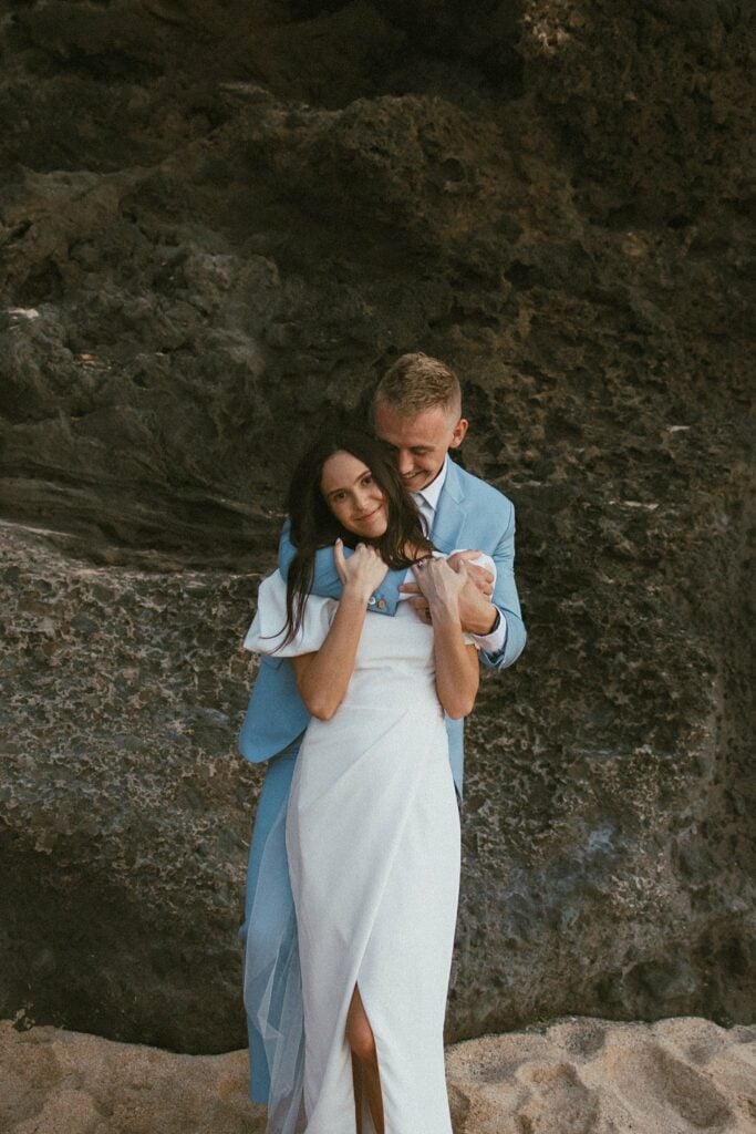 the groom is hugging the bride from the behind while they stand in front of a cliff on the beach