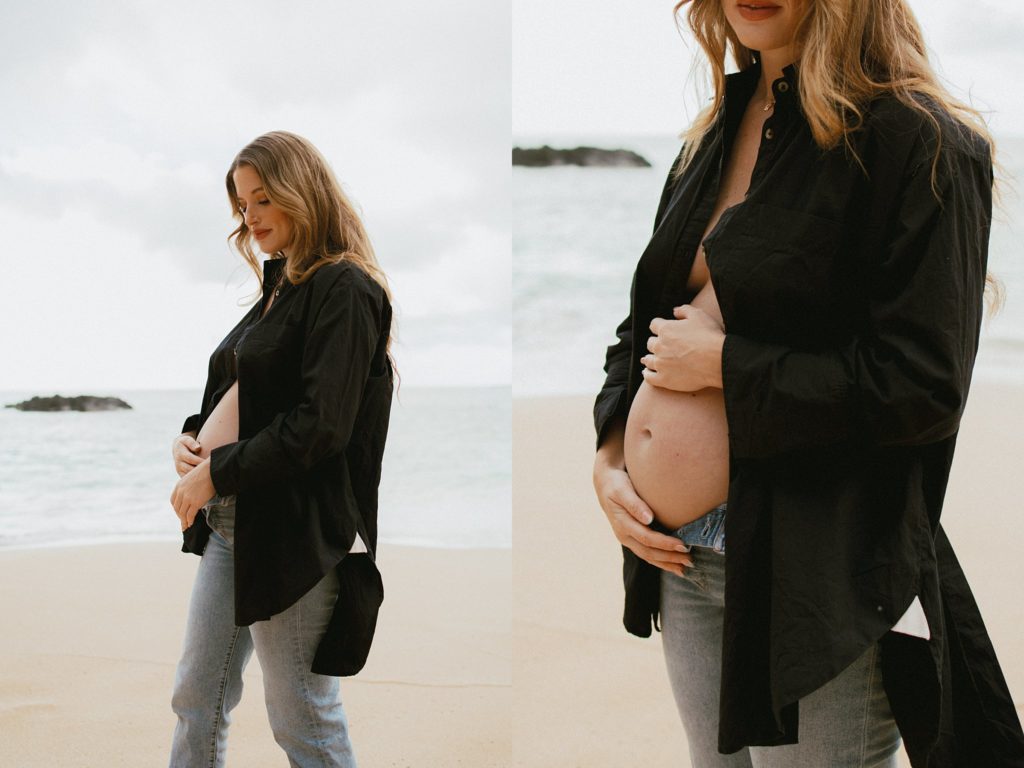 mother is holding her pregnant belly on the beach while wearing a black, unbuttoned, button down shirt 
