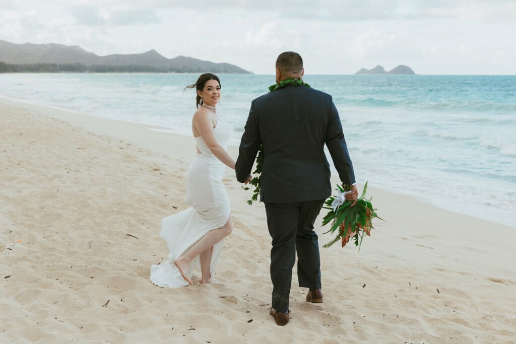 bride and groom are holding hands walking along the beach, bride is looking back and smilling at the groom