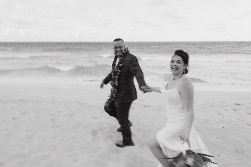 black and white shot of bride and groom running along the beach and laughing together
