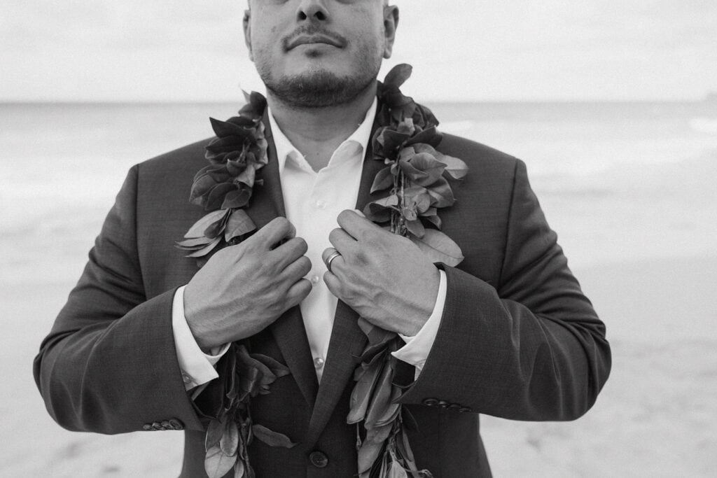 black and white shot of the groom fixing his jacket with a lei wrappeda around his neck