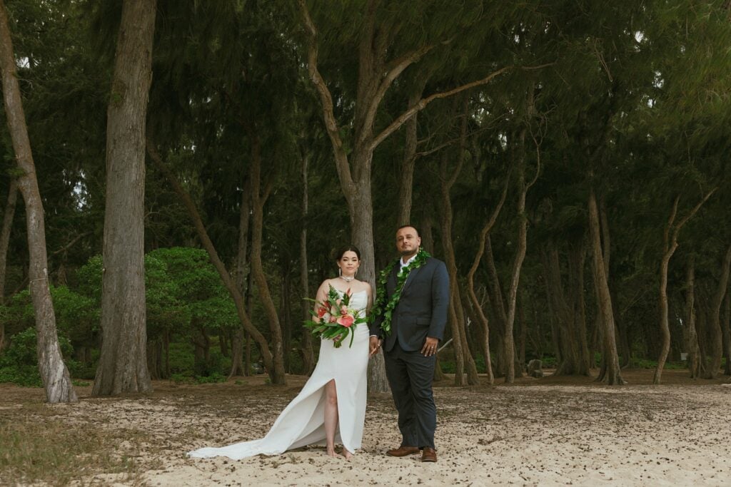 bride and groom are standing on the beach, encircled by trees and looking at the camera seriously