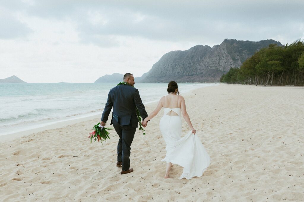 bride and groom are holding hands and walking along the beach with mountain scenery in the back