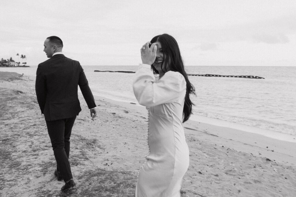 black and white photo of hao walking along the beach while julie looks back and smiles at the camera