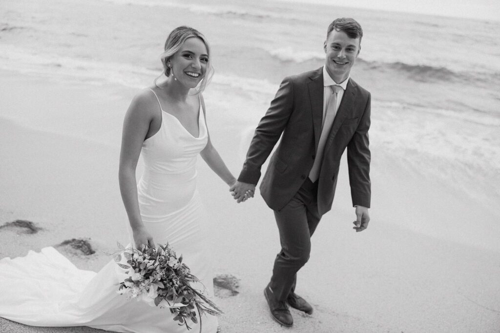 black and white shot of bride and groom holding hands walking along the beach