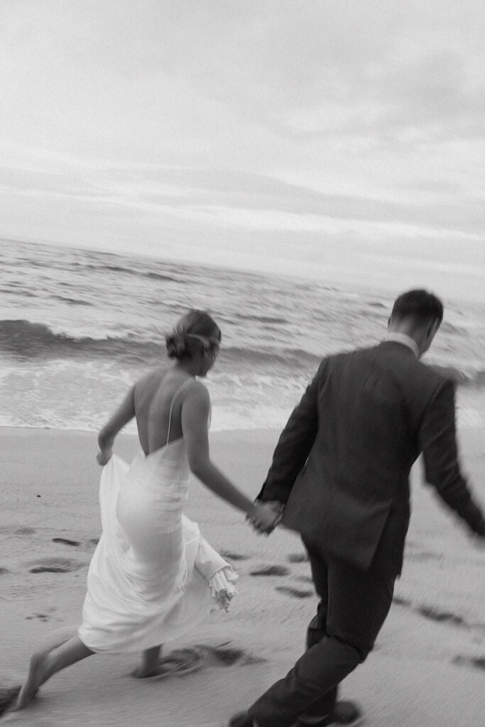 black and white shot of bride and groom running along the beach while holding hands