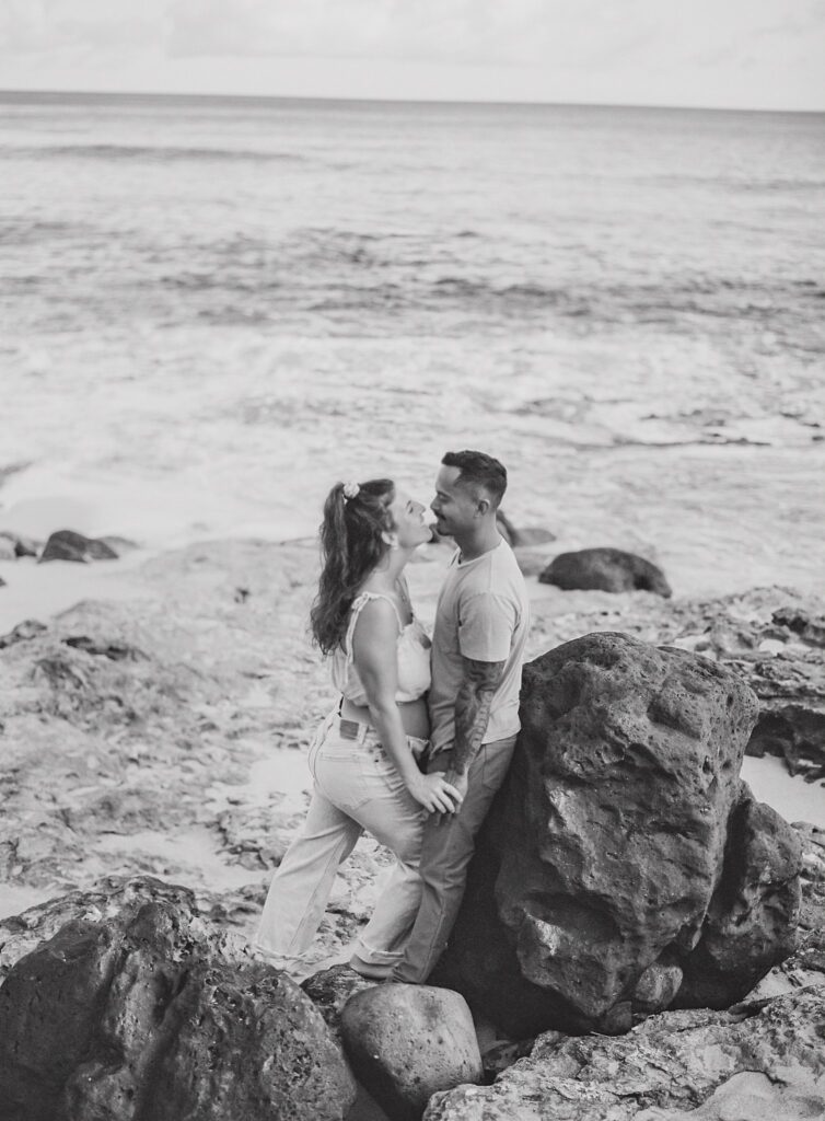 black and white film photo of couple leaning against a rock on the beach during their maternity photoshoot